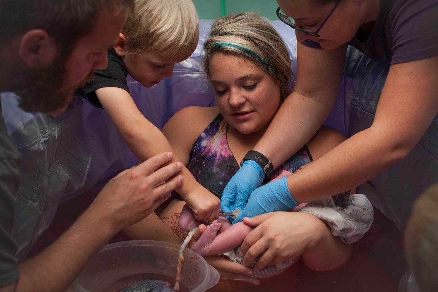 A family participates in a water birth at home, guided by a licensed midwife in Indiana, with a young sibling assisting in cutting the umbilical cord.