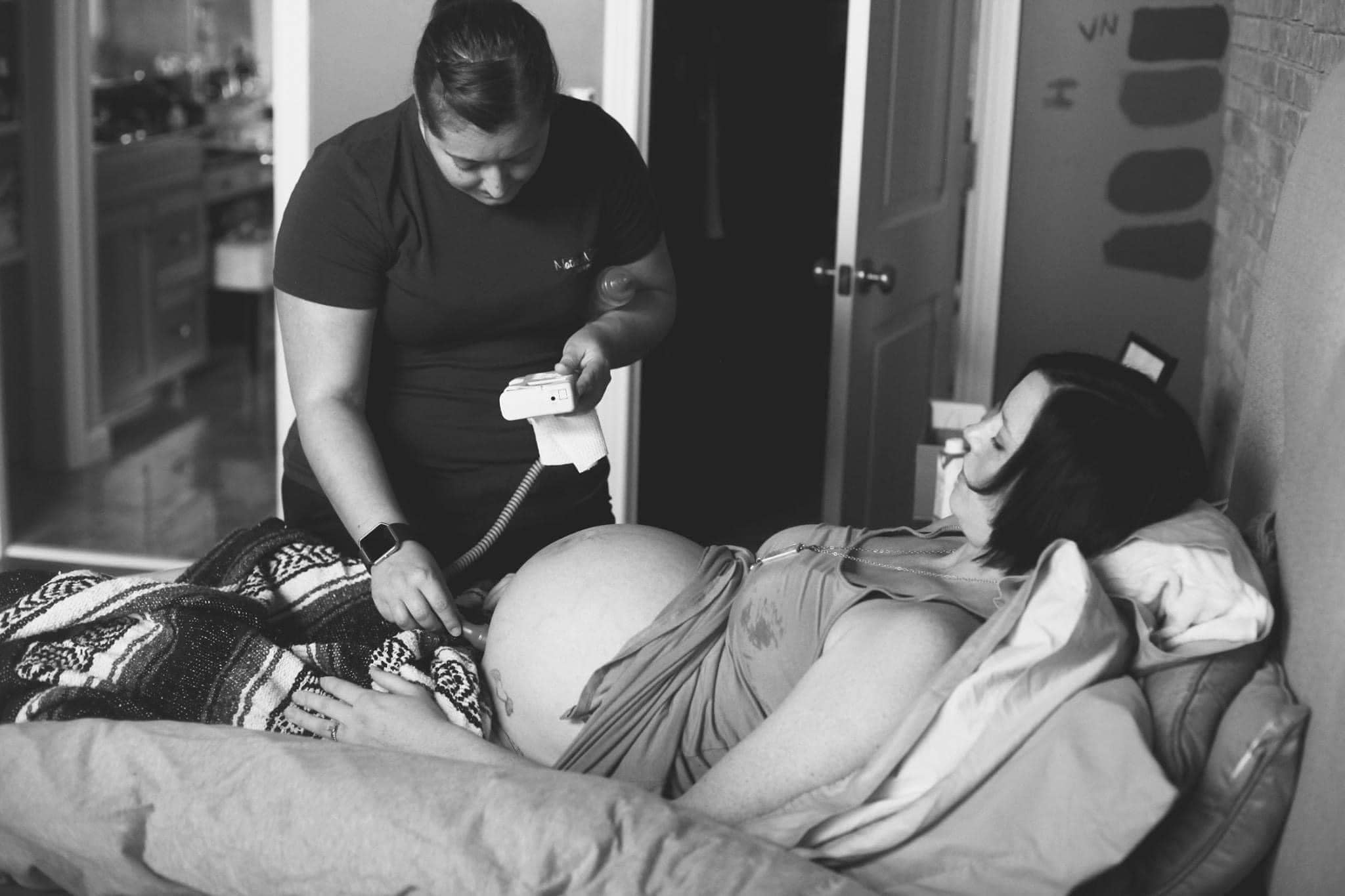 Midwife monitoring fetal heart tones using a Doppler device on a pregnant woman lying in bed.