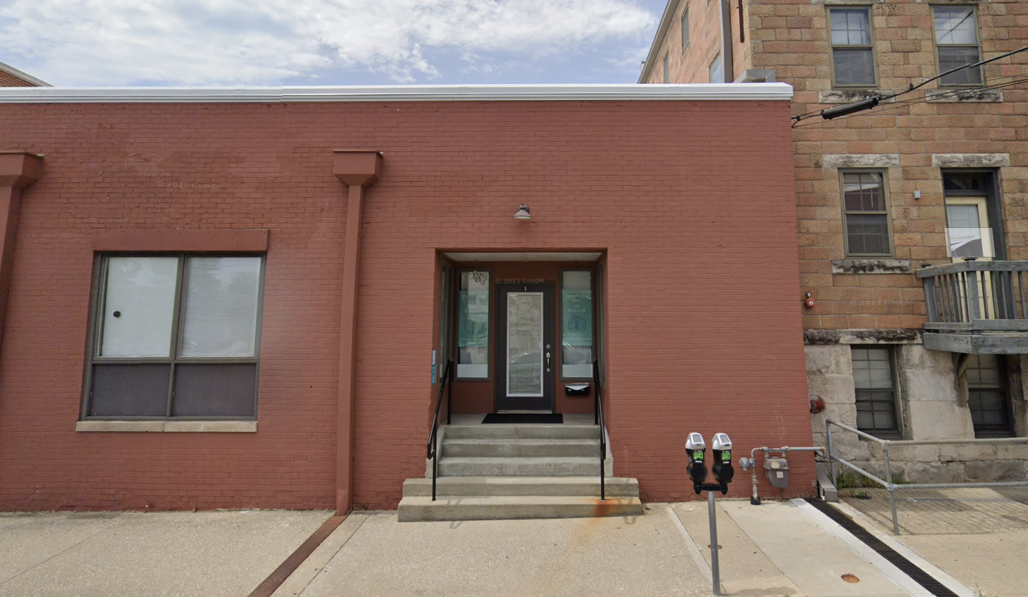 Entrance to Mother Nurtured Midwifery in Bloomington, showing a brick building with a glass door, steps leading up to the entrance, and two parking meters in front.